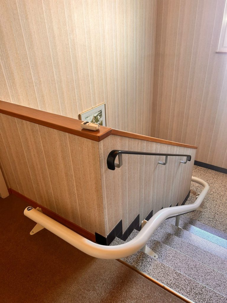 LSM Mobility Siège monte-escaliers blanc installé dans une cage d'escalier avec moquette marron au départ et sol en terrazzo. Rampe noire et mur tapissé de beige. Confort et accessibilité dans le Gros-de-Vaud.
