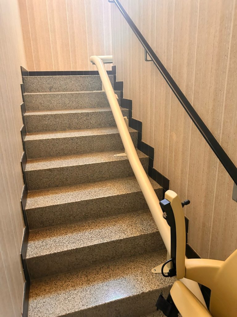 LSM Mobility Escalier intérieur avec un siège monte-escaliers beige installé. L'escalier est en granit gris avec des bordures noires. Une rampe noire est fixée au mur. Idéal pour un article sur un siège monte-escaliers dans le Gros-de-Vaud.