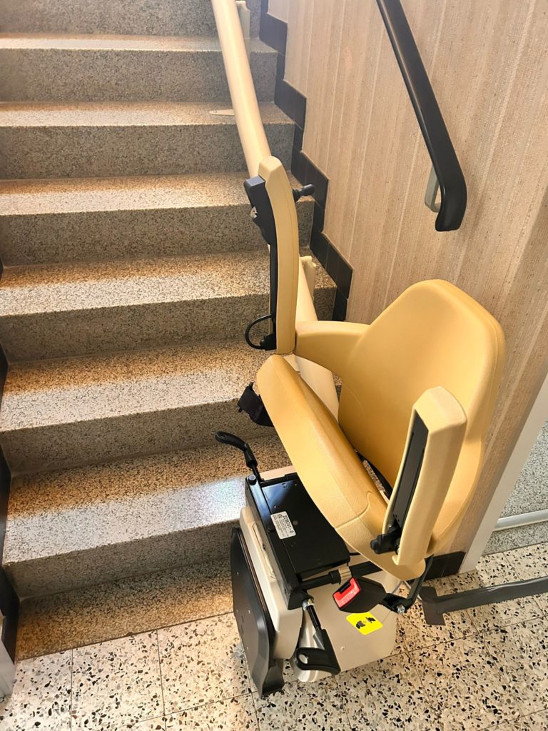 LSM Mobility Siège monte-escaliers beige installé dans un immeuble du Gros-de-Vaud, offrant une solution d'accessibilité pratique et sécurisée. L'escalier est en béton avec des marches grises.