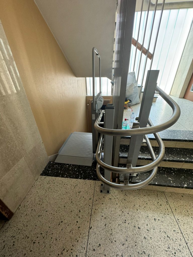 LSM Mobility Plateforme monte-escaliers Ascendor PLK8 installée dans une cage d'escalier lumineuse à Neuchâtel. La plateforme gris clair est repliée, prête à l'emploi. Rampe en inox et sol en terrazzo moucheté.