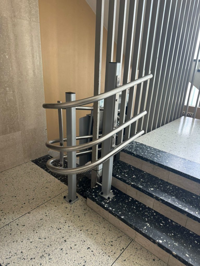 LSM Mobility Plateforme monte-escaliers Ascendor PLK8 installée sur un escalier intérieur avec des marches en terrazzo noir et blanc. La plateforme en métal argenté est repliée, prête à l'utilisation.