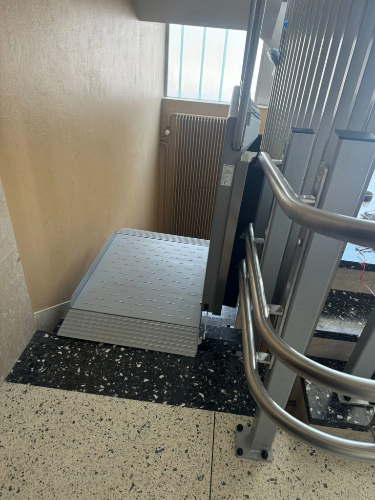 LSM Mobility Plateforme monte-escalier Ascendor PLK8 repliée dans un escalier à Neuchâtel. L'appareil gris est rangé contre le mur, avec sa plateforme grise pliée. Sol en terrazzo noir et blanc.