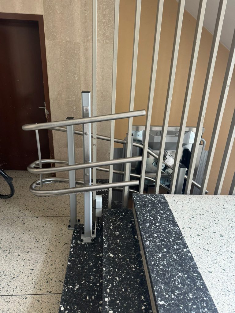 LSM Mobility Plateforme monte-escaliers Ascendor PLK8 installée à Neuchâtel. Vue d'un monte-escalier avec plateforme, monté près d'un escalier aux barreaux verticaux et d'une porte marron.