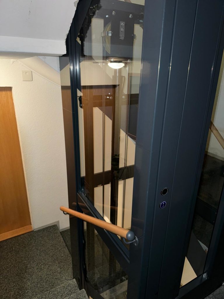 LSM Mobility Homelift moderne en verre et métal gris installé dans une cage d'escalier. L'ascenseur résidentiel facilite l'accès aux étages et s'intègre parfaitement à l'architecture intérieure.