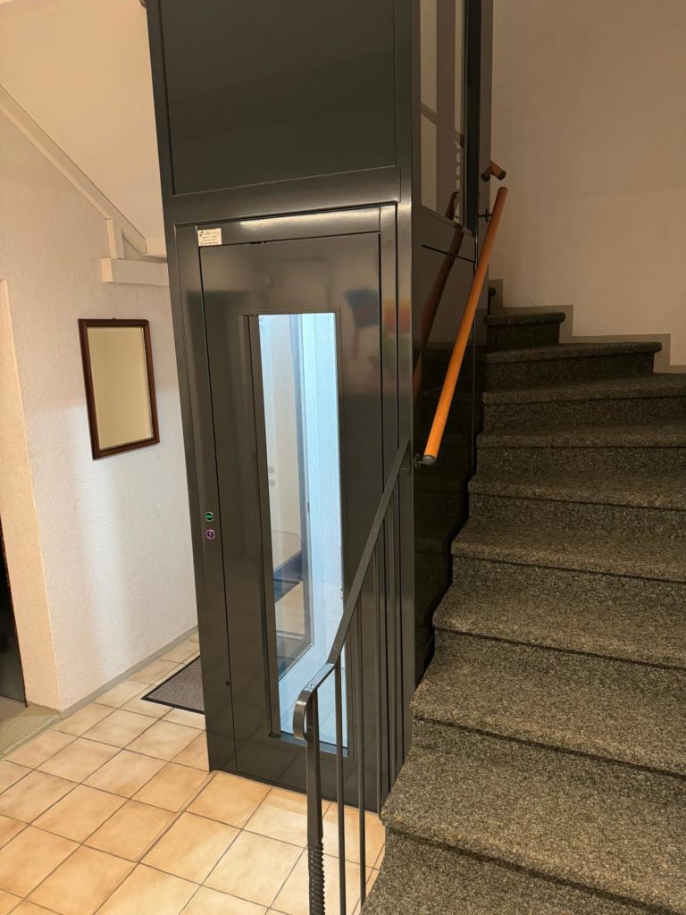 LSM Mobility Ascenseur résidentiel gris moderne installé dans une cage d'escalier avec marches en moquette. Un homelift qui facilite l'accès aux étages sur le littoral neuchâtelois.