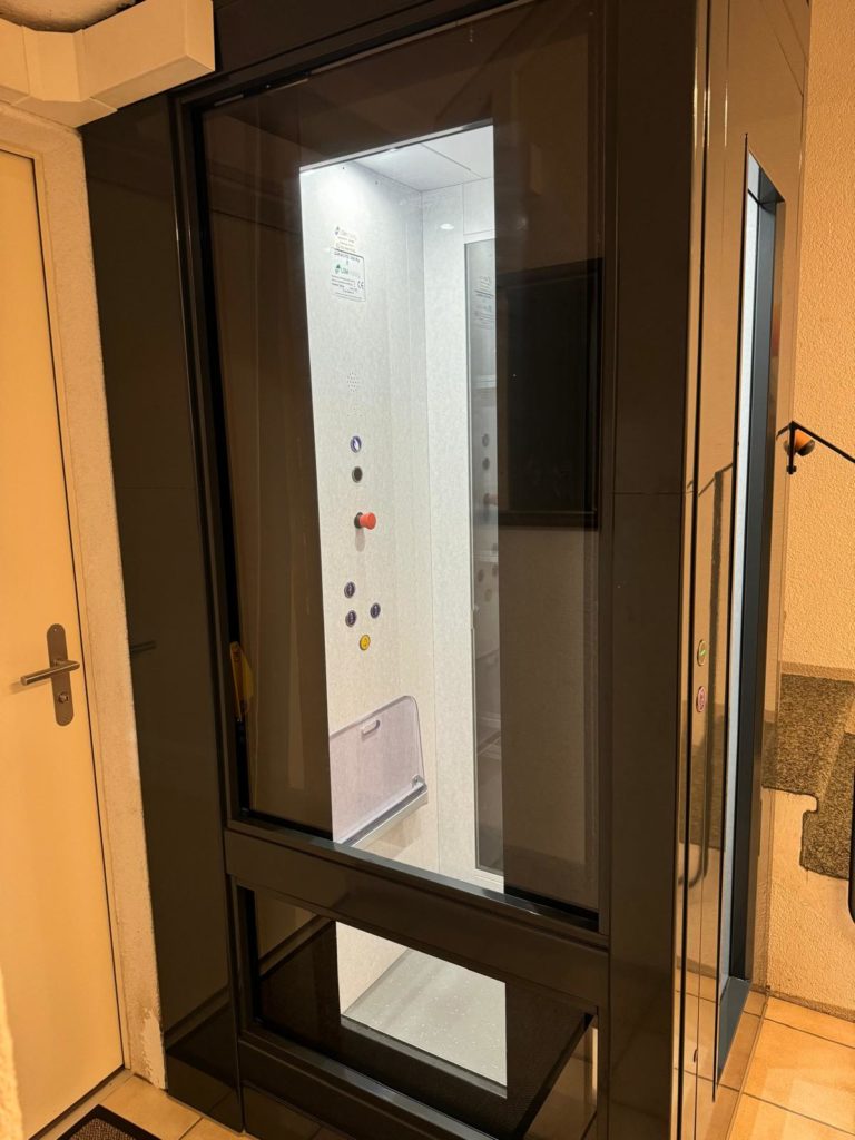 LSM Mobility Homelift moderne avec cadre noir et parois vitrées, installé près d'une porte blanche. Intérieur clair avec boutons de commande. Solution d'accessibilité élégante sur le littoral neuchâtelois.