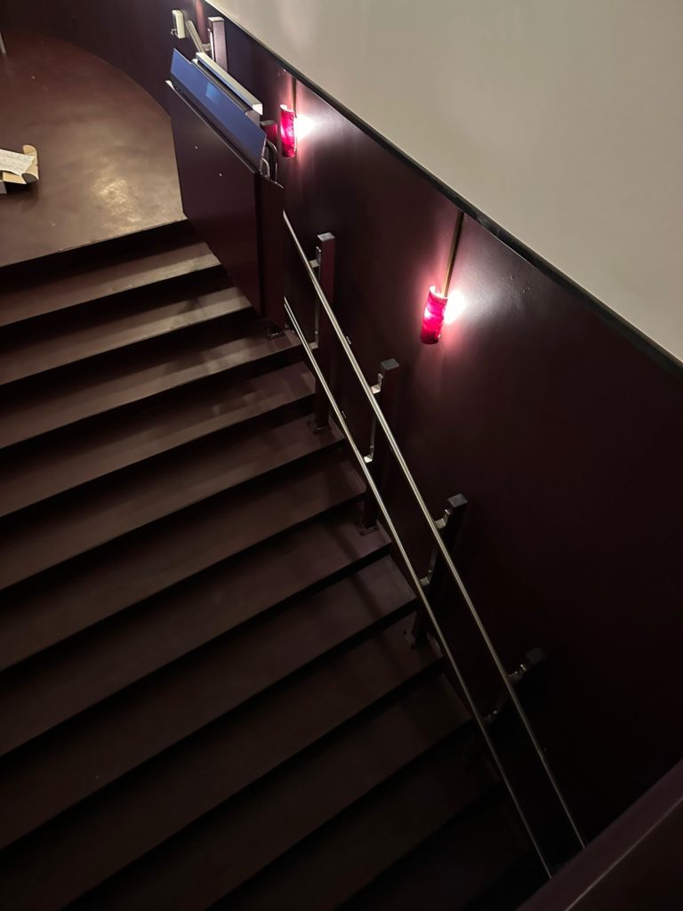 LSM Mobility Escalier descendant au Cinéma Korso à Fribourg, éclairé par des lumières rouges. Rampe d'accès en métal sur le côté. Plateforme monte-escaliers potentielle.