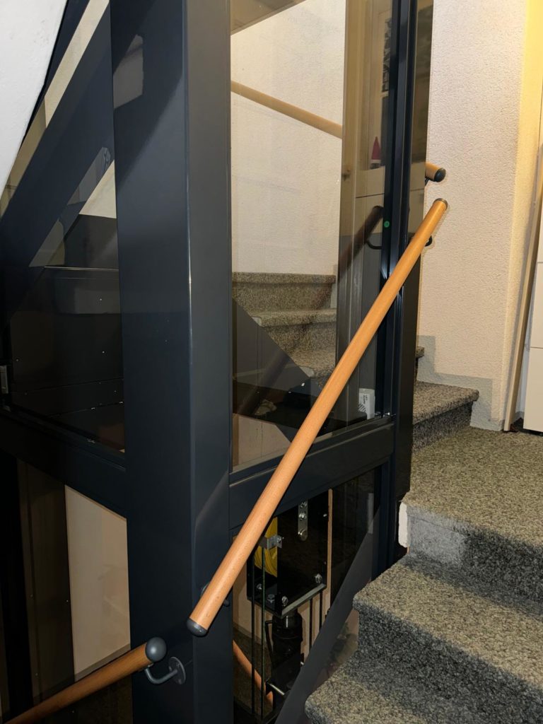 LSM Mobility Intérieur avec homelift moderne près d'un escalier en pierre. La structure métallique anthracite contraste avec la rampe en bois clair. Idéal pour une maison sur le littoral neuchâtelois.
