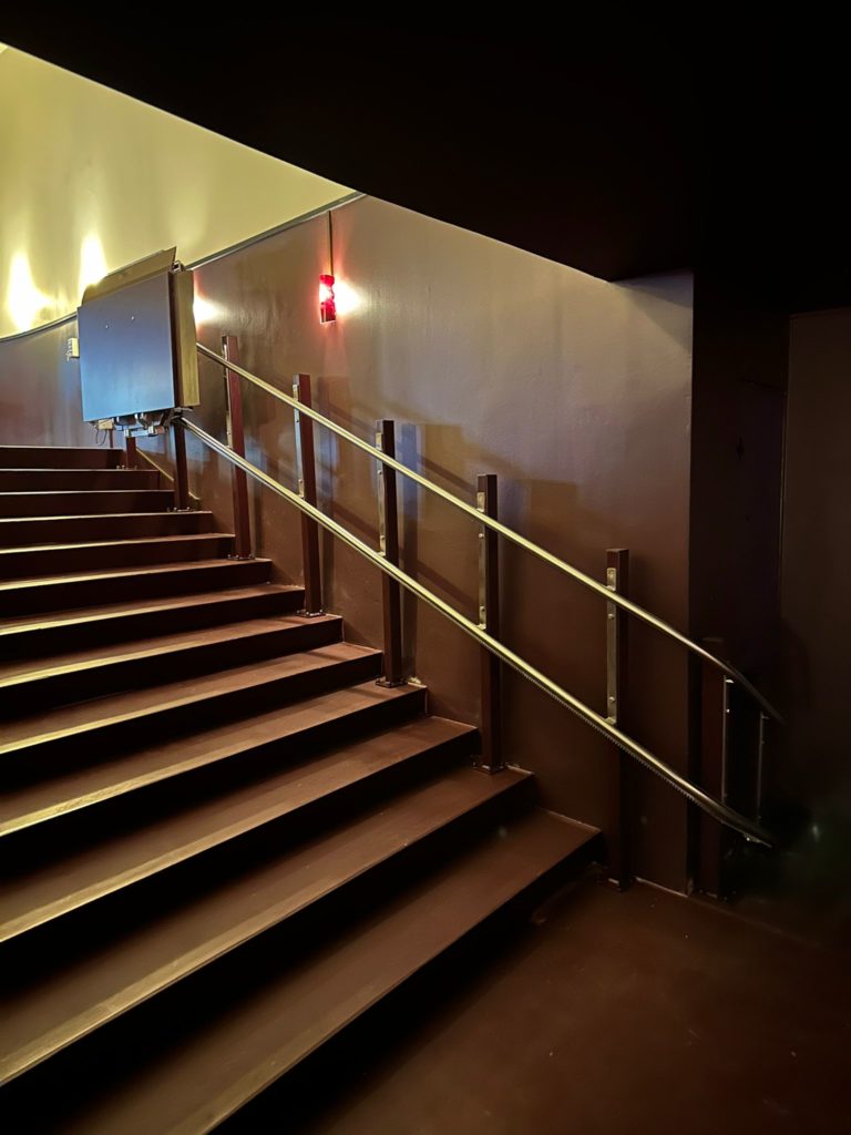LSM Mobility Escalier marron avec plateforme monte-escaliers installée au Cinéma Korso à Fribourg. Rampe en métal et éclairage rouge.