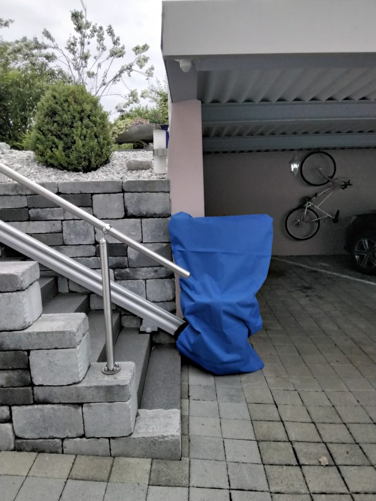 LSM Mobility Siège monte-escaliers extérieur protégé par une housse bleue, près d'un escalier en pierre et d'un carport. Un vélo est accroché au mur du carport.