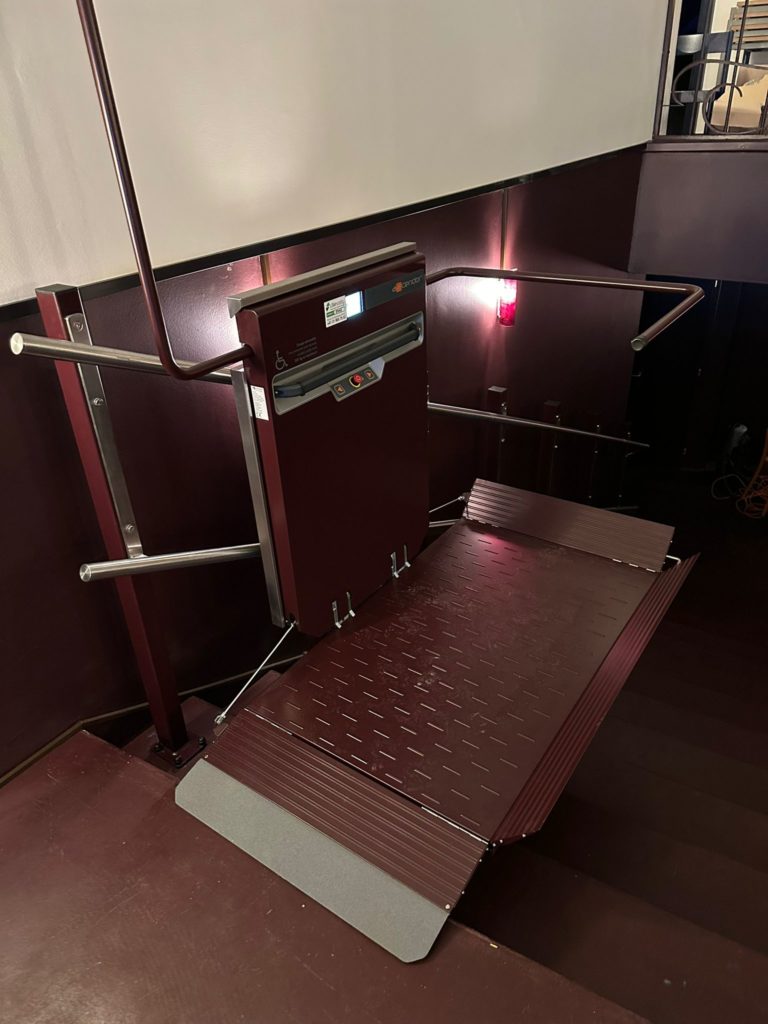 LSM Mobility Plateforme monte-escaliers Cinéma Korso à Fribourg, marron et gris, installée dans un escalier. Rampe d'accès pour fauteuils roulants. Ascenseur pour personnes à mobilité réduite.