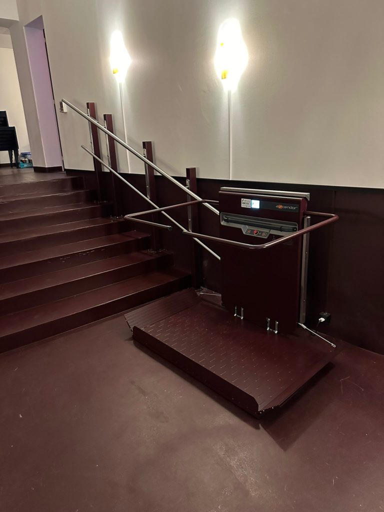LSM Mobility Plateforme monte-escaliers Ascendor rouge bordeaux installée au Cinéma Korso à Fribourg. Rampe d'accès et escaliers assortis, murs blancs éclairés par des appliques modernes.
