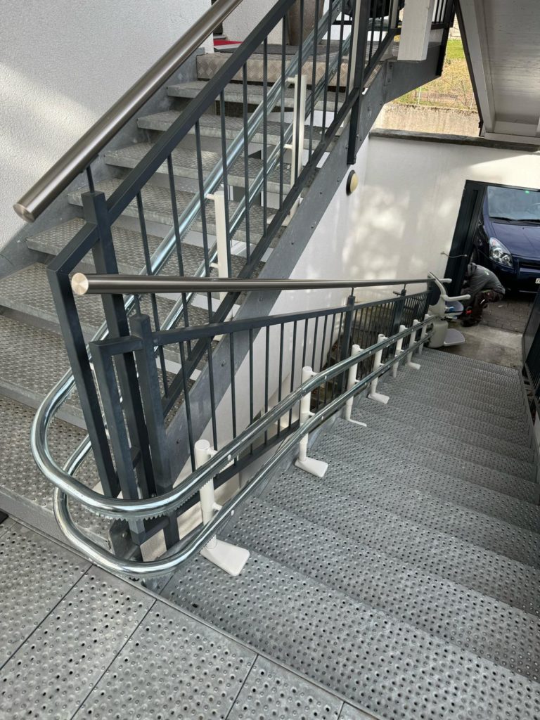 LSM Mobility Siège monte-escalier extérieur Handicare 4000 installé sur un escalier métallique menant à une entrée. Le dispositif est équipé d'une rampe de sécurité argentée.