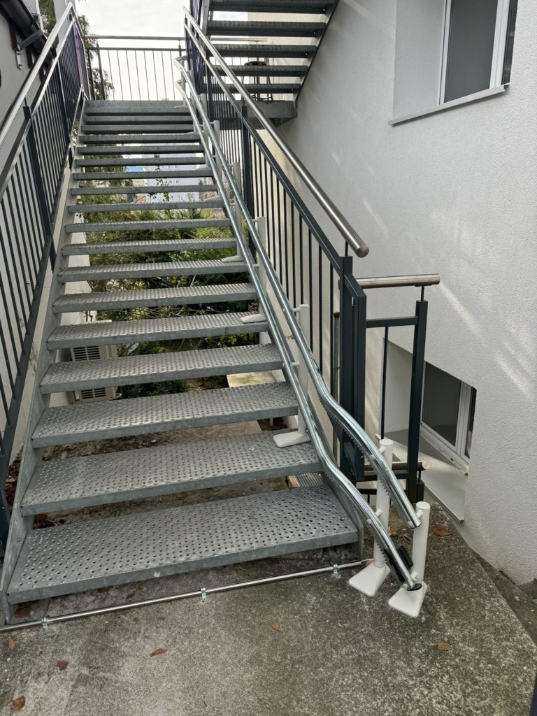 LSM Mobility Escalier extérieur en métal gris montant vers un bâtiment blanc. Le siège monte-escaliers Handicare 4000 est visible en haut des marches.