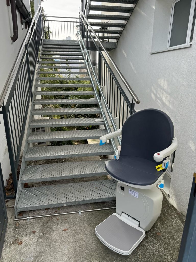 LSM Mobility Siège monte-escaliers extérieur Handicare 4000 installé sur un escalier métallique gris à l'extérieur d'un bâtiment blanc. Le siège est replié, prêt à l'emploi.