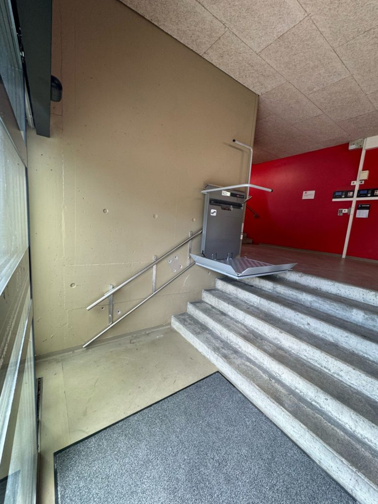 LSM Mobility Plateforme élévatrice grise installée sur un escalier en béton clair, avec des rampes métalliques. Mur beige en arrière-plan, partie rouge visible. Remplacement plateforme Stade de la Maladière.