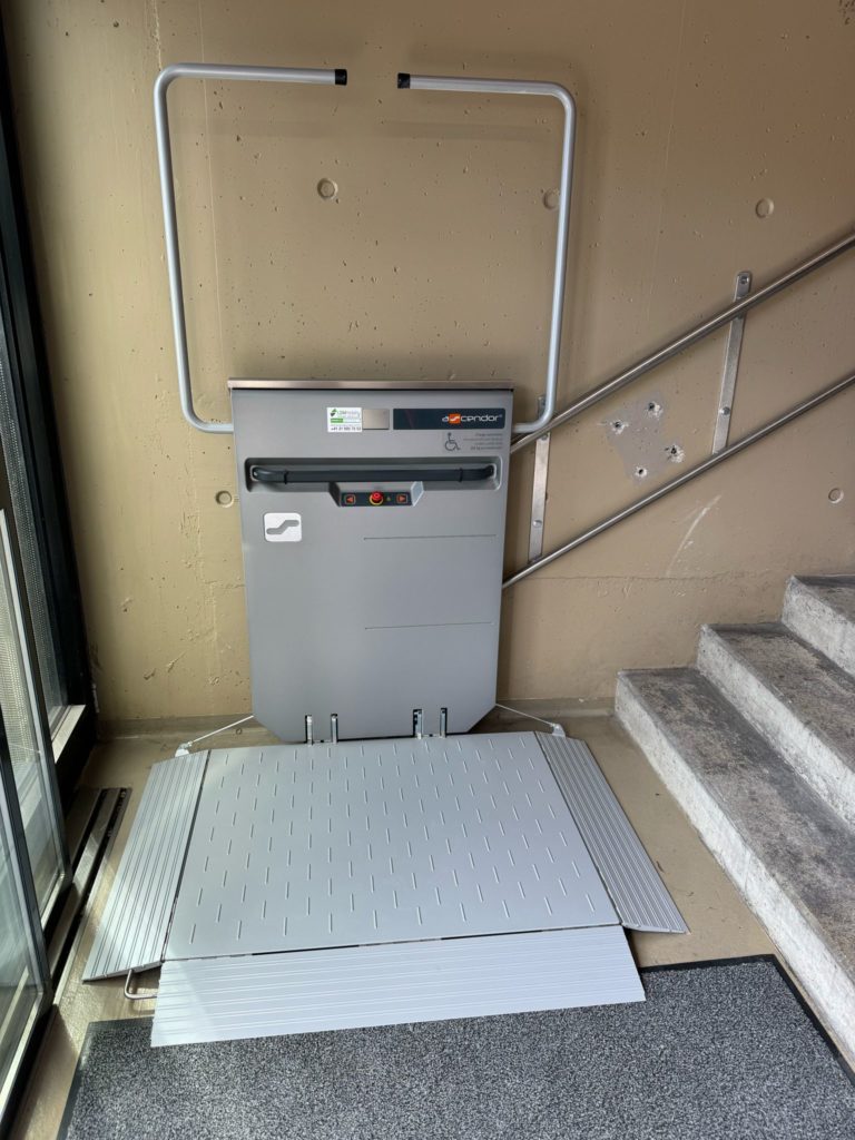 LSM Mobility Plateforme élévatrice gris clair installée à côté d'un escalier en béton dans un bâtiment. Rampe déployée. Remplacement d'une plateforme au stade de la Maladière à Neuchâtel.