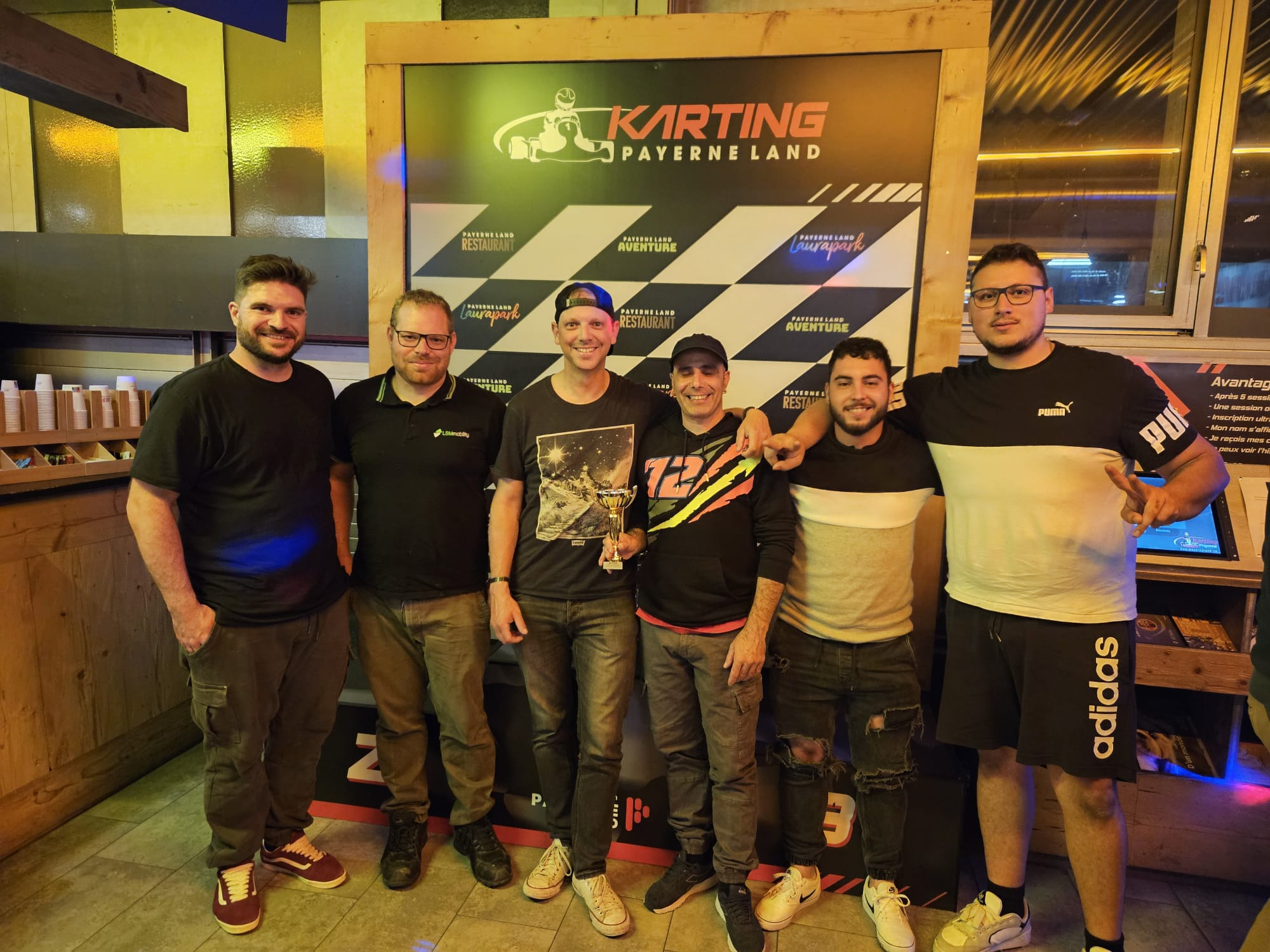 LSM Mobility Une équipe pose fièrement devant le logo Karting Payerne Land après la Coupe interentreprise 2025. On voit six hommes souriants, dont un tenant un trophée.