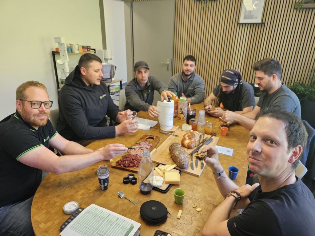 LSM Mobility L'équipe partage un brunch convivial après une séance de coordination. Hommes autour d'une table ronde avec charcuterie, fromage, pain et jus. Ambiance décontractée et collaborative.
