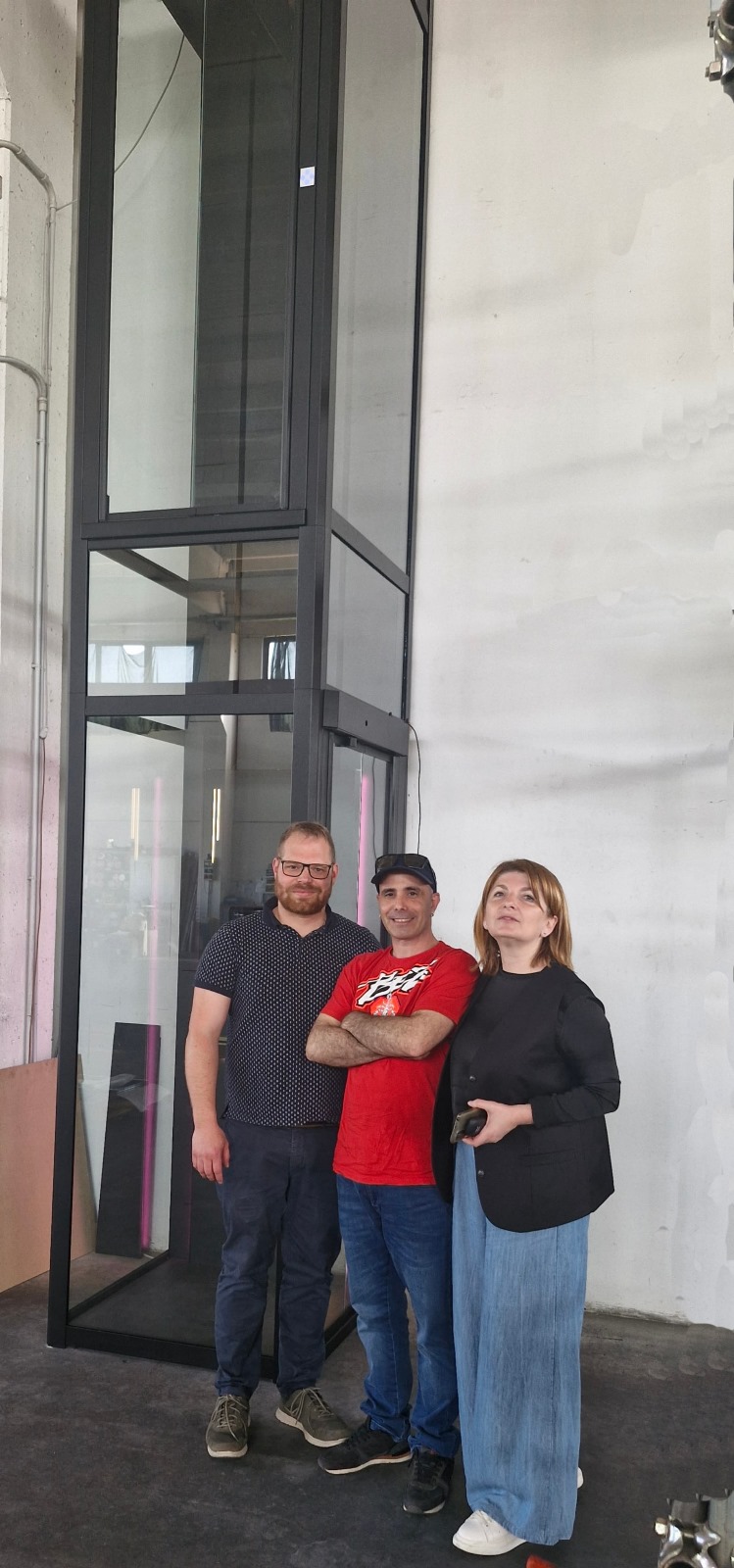 LSM Mobility Trois personnes sourient devant un ascenseur résidentiel moderne de Nova Elevators, lors d'une visite. L'ascenseur a une structure noire et des parois en verre.