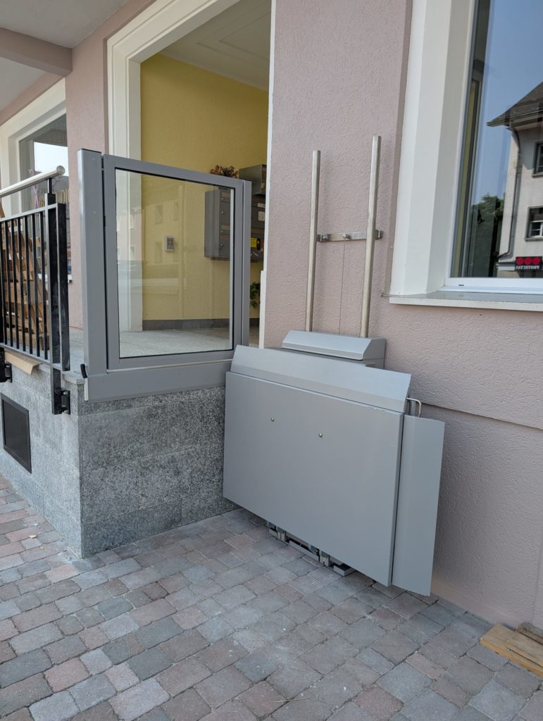 LSM Mobility Plateforme élévatrice Ascendor PLV9 grise installée à l'extérieur d'un bâtiment, offrant accessibilité. La plateforme est repliée, montrant son design compact et sa facilité d'intégration architecturale.