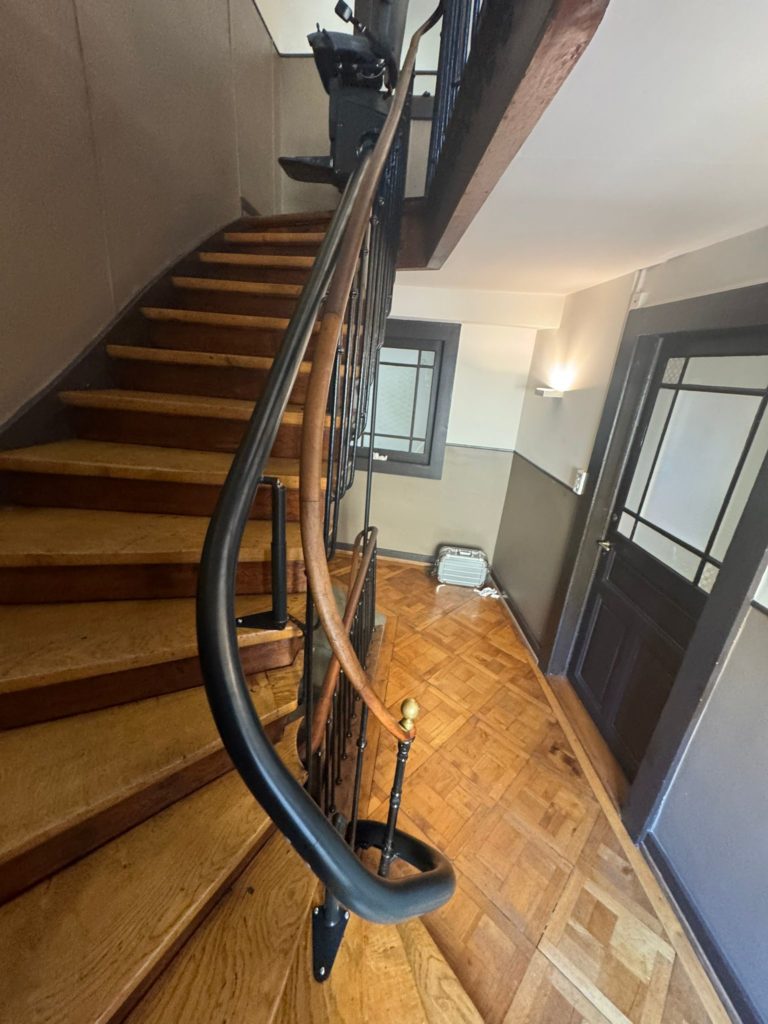 LSM Mobility Vue plongeante sur un escalier en bois avec rampe en fer forgé. Un siège monte-escaliers Handicare monorail personnalisé est installé. Parquet en chevrons et porte sombre visible.