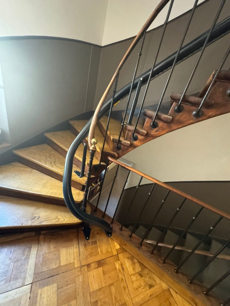 LSM Mobility Escalier en bois tournant avec rampe en fer forgé et parquet en damier. Illustration possible d'un siège monte-escaliers Handicare monorail personnalisé.