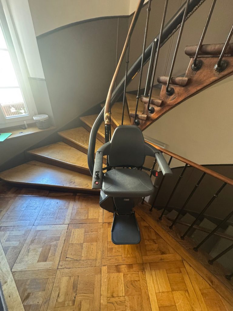 LSM Mobility Siège monte-escalier Handicare monorail personnalisé installé sur un escalier en bois tournant avec une rampe en fer forgé. Le siège est gris foncé et l'escalier est éclairé par une fenêtre.