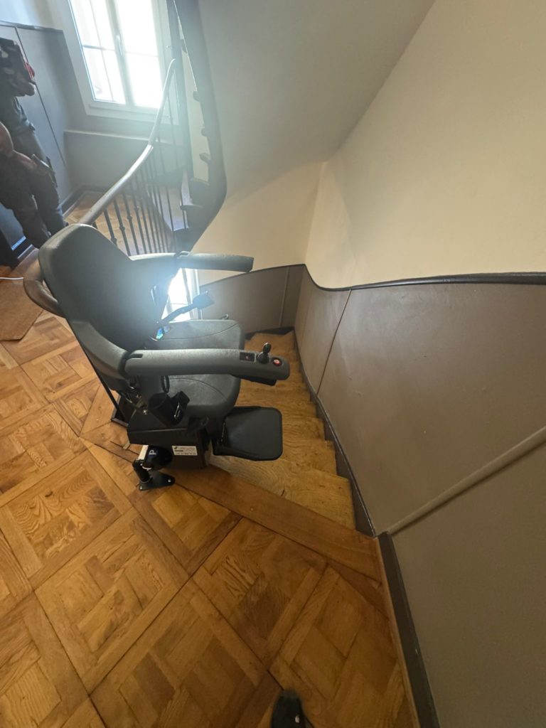 LSM Mobility Un siège monte-escaliers Handicare monorail personnalisé installé sur un escalier en bois dans une maison. Le siège est noir, avec des accoudoirs et un repose-pieds.