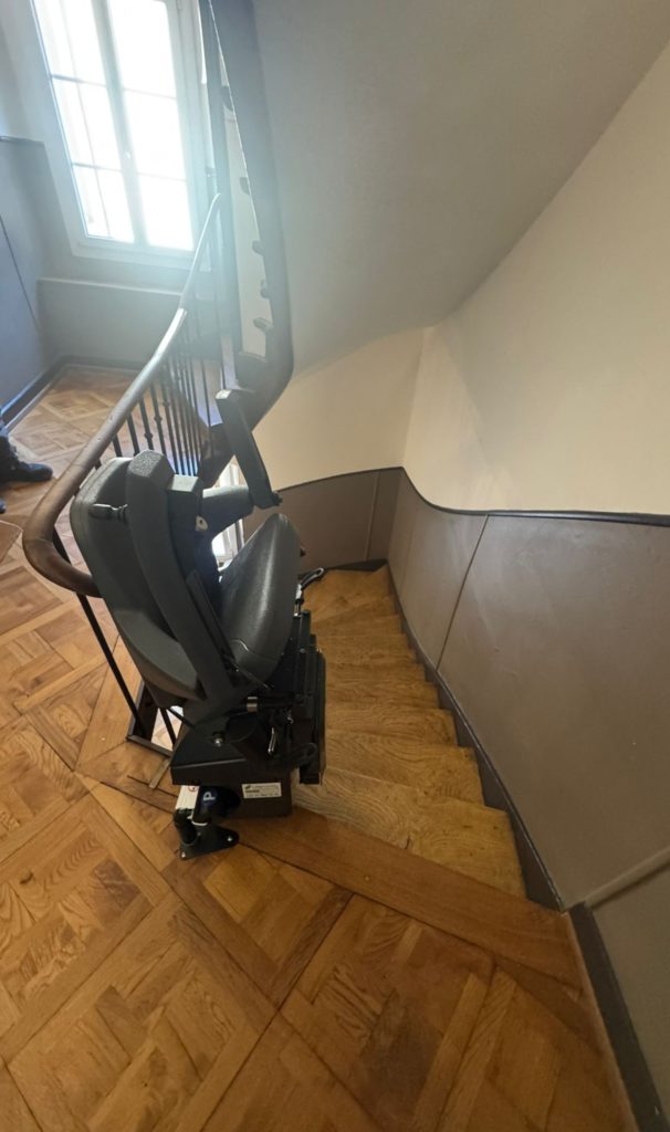 LSM Mobility Siège monte-escaliers Handicare monorail gris installé dans un escalier en bois clair avec rampe en fer forgé. La chaise est repliée, prête à l'emploi.