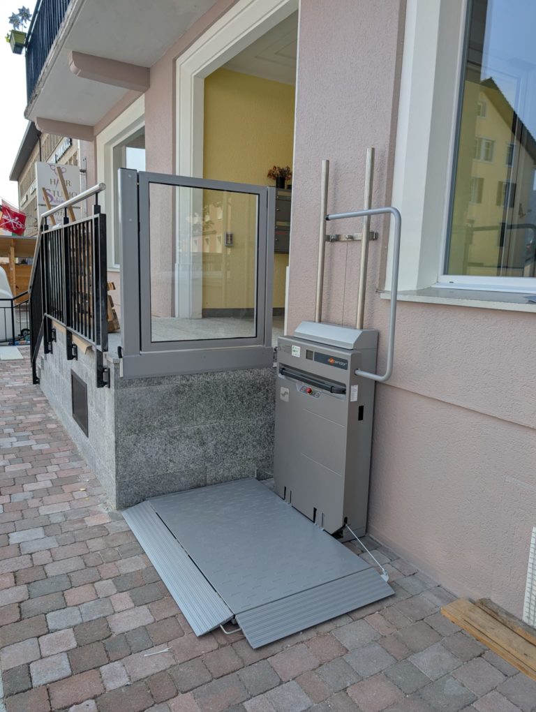 LSM Mobility Plateforme élévatrice Ascendor PLV9 grise installée à l'extérieur d'un bâtiment rose pour l'accessibilité PMR. Rampe d'accès en métal et structure sécurisée. Ascendor visible.
