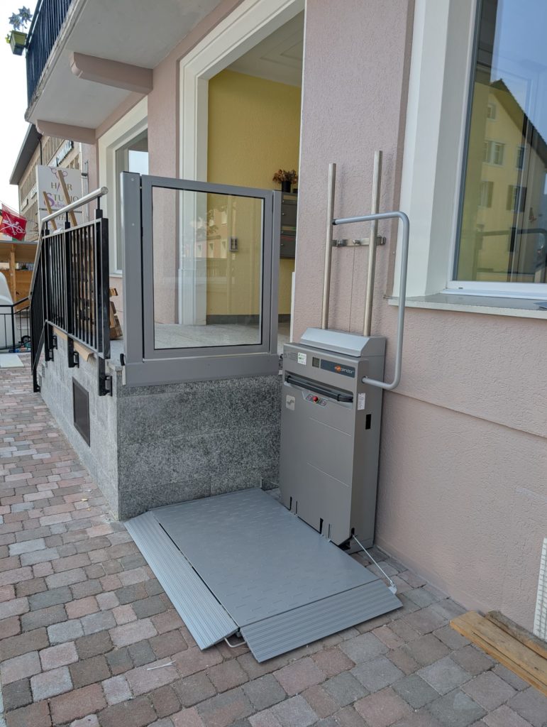 LSM Mobility Plateforme élévatrice Ascendor PLV9 grise installée à l'extérieur d'un bâtiment rose pour l'accessibilité PMR. Rampe d'accès en métal et structure sécurisée. Ascendor visible.