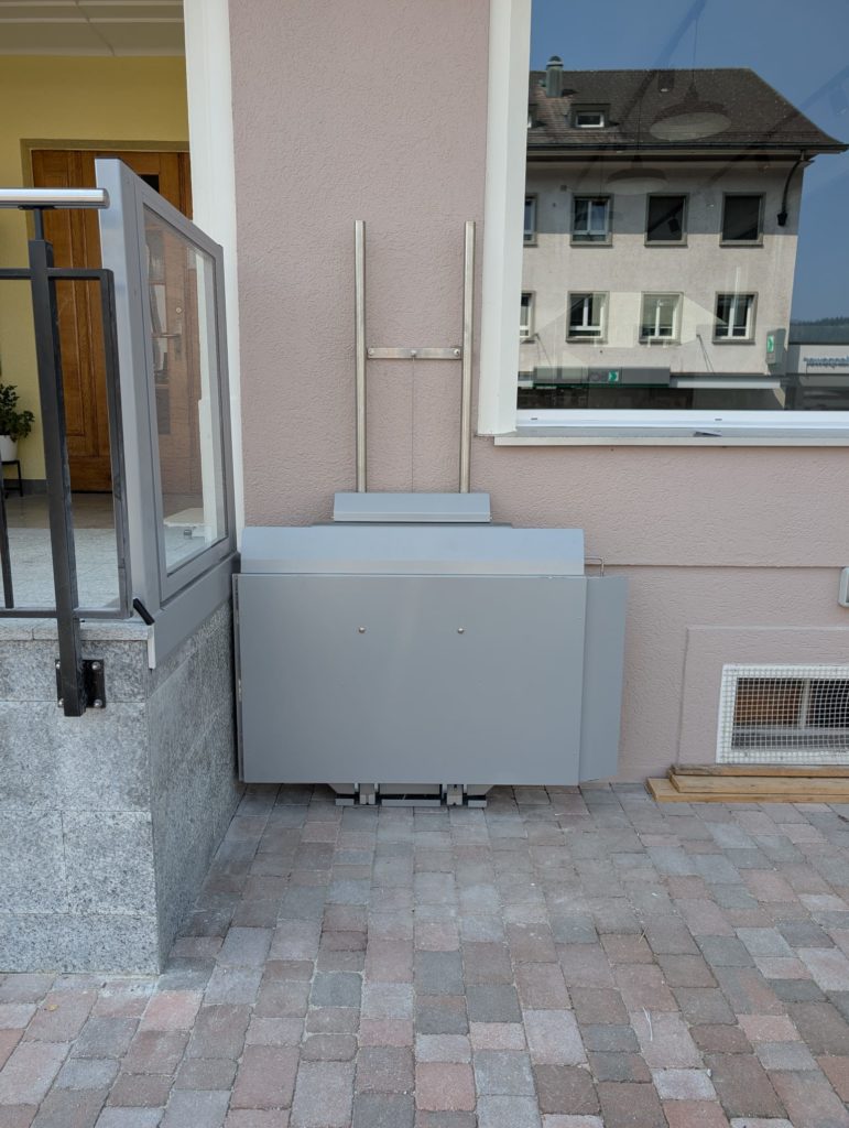 LSM Mobility Plateforme élévatrice Ascendor PLV9 gris clair installée à l'extérieur d'un bâtiment beige, avec une porte d'entrée et une fenêtre reflétant un autre bâtiment. Accès facilité pour personnes à mobilité réduite.