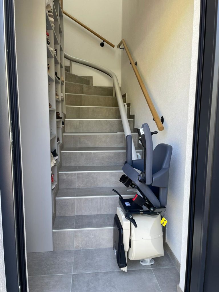 LSM Mobility Monte-escalier moderne gris clair installé dans un escalier carrelé gris. Rail incurvé pour suivre la forme de l'escalier. Placard à chaussures intégré à gauche.