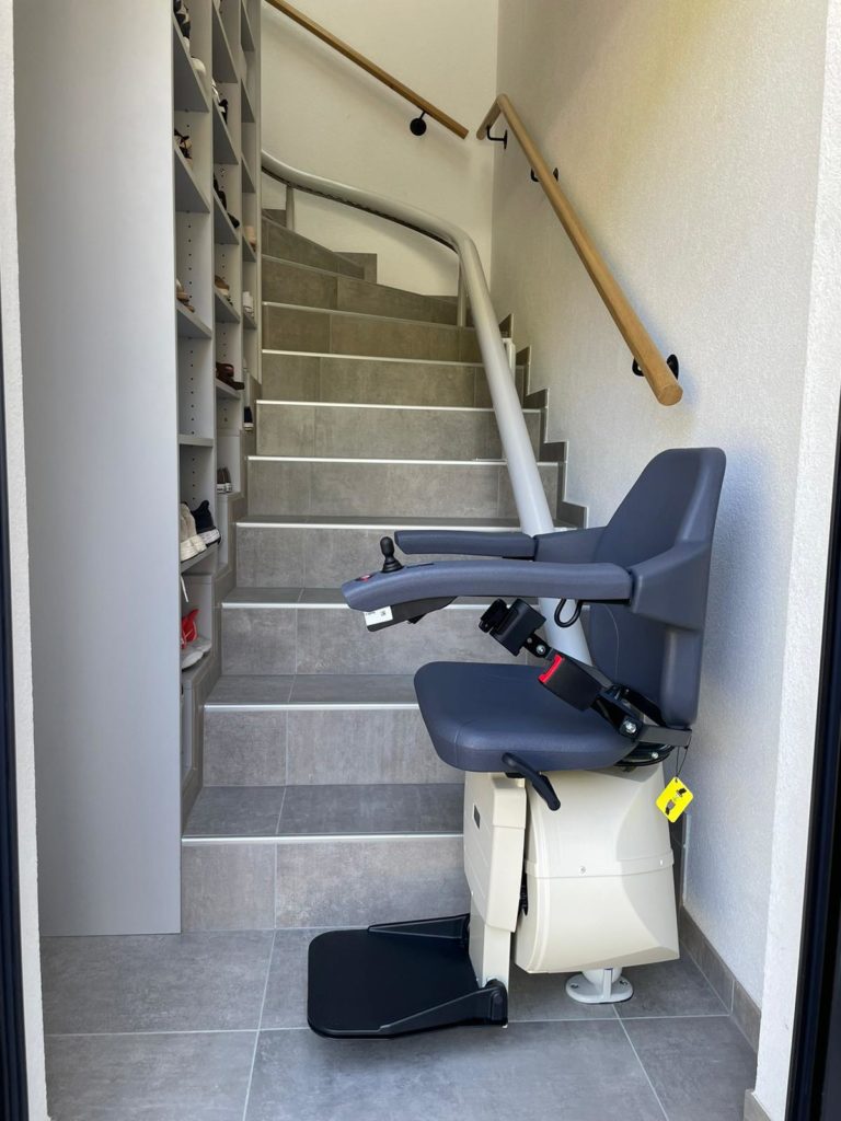 LSM Mobility Monte-escalier gris moderne installé sur un escalier intérieur carrelé avec rampe en bois. Un placard à chaussures intégré est visible sur le côté. Solution d'accessibilité pour maison.