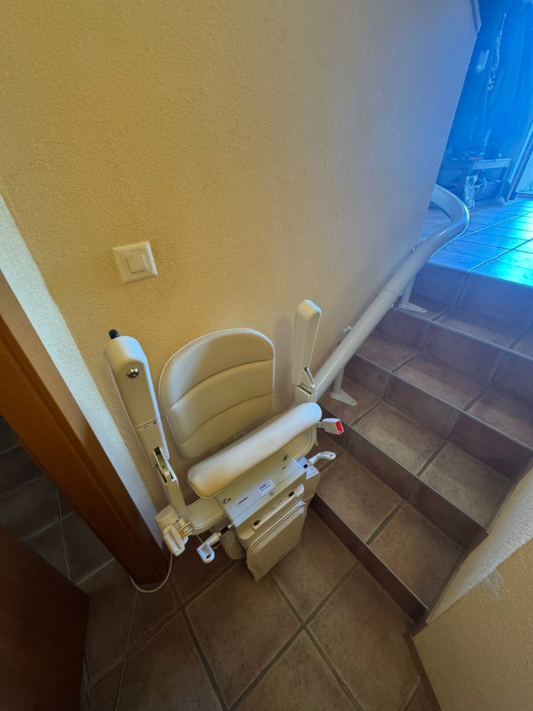 LSM Mobility Monte-escalier blanc installé le long d'un mur beige dans un escalier intérieur avec des marches carrelées marron. Solution d'accessibilité domestique.