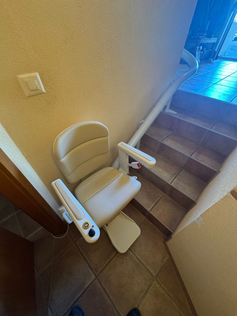 LSM Mobility Monte-escalier beige installé dans un escalier carrelé marron. Le siège est replié contre le mur, avec une rampe blanche qui suit la courbe de l'escalier.