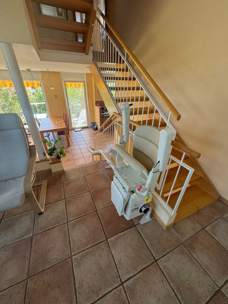 LSM Mobility Un monte-escalier beige installé sur un escalier en bois clair dans une maison avec carrelage en terre cuite. Vue sur une salle à manger lumineuse.