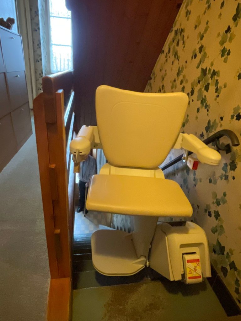 LSM Mobility Monte-escalier beige installé dans un intérieur avec papier peint floral. Un homme se tient en bas des marches. Confort et accessibilité à domicile.