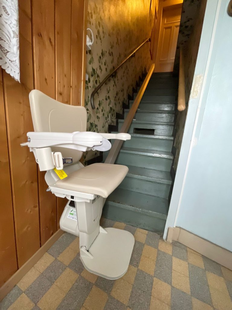 LSM Mobility Siège monte-escaliers Handicare 1100 beige installé dans un escalier étroit avec papier peint floral et rampe en métal. L'escalier est peint en gris-vert et le sol est carrelé.