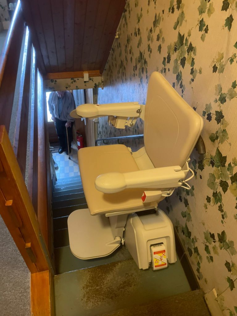 LSM Mobility Monte-escalier beige installé dans un couloir tapissé de motifs floraux. Un homme âgé se tient en haut des escaliers, offrant un aperçu d'une maison lumineuse.