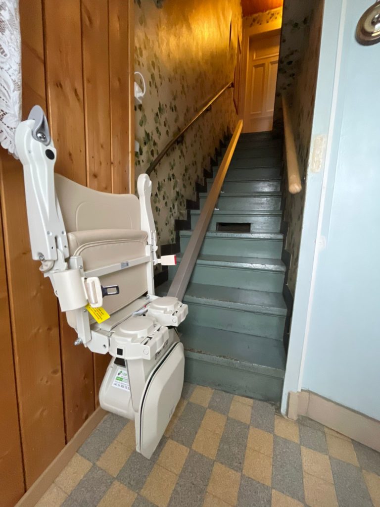 LSM Mobility Un monte-escalier beige replié contre un mur en bois, en bas d'un escalier étroit aux marches vertes et rampe métallique. Papier peint floral vintage sur le mur de l'escalier.