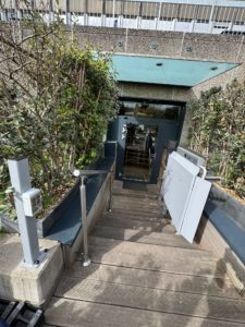 LSM Mobility Plateforme élévatrice PLG7 extérieure menant à une entrée de bâtiment moderne. La plateforme est installée le long d'un escalier en bois, bordé de végétation.