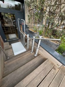 LSM Mobility Plateforme élévatrice PLG7 extérieure pour fauteuil roulant, installée sur des marches en bois devant une entrée de bâtiment moderne. L'accès facilité pour PMR est mis en avant.