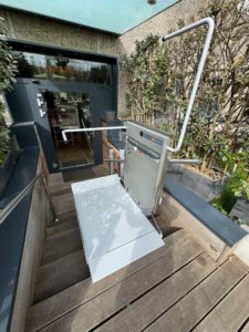 LSM Mobility Plateforme élévatrice PLG7 installée sur des marches extérieures menant à une porte d'entrée vitrée. L'élévateur gris clair est doté d'une rampe et d'une barre de sécurité en acier.