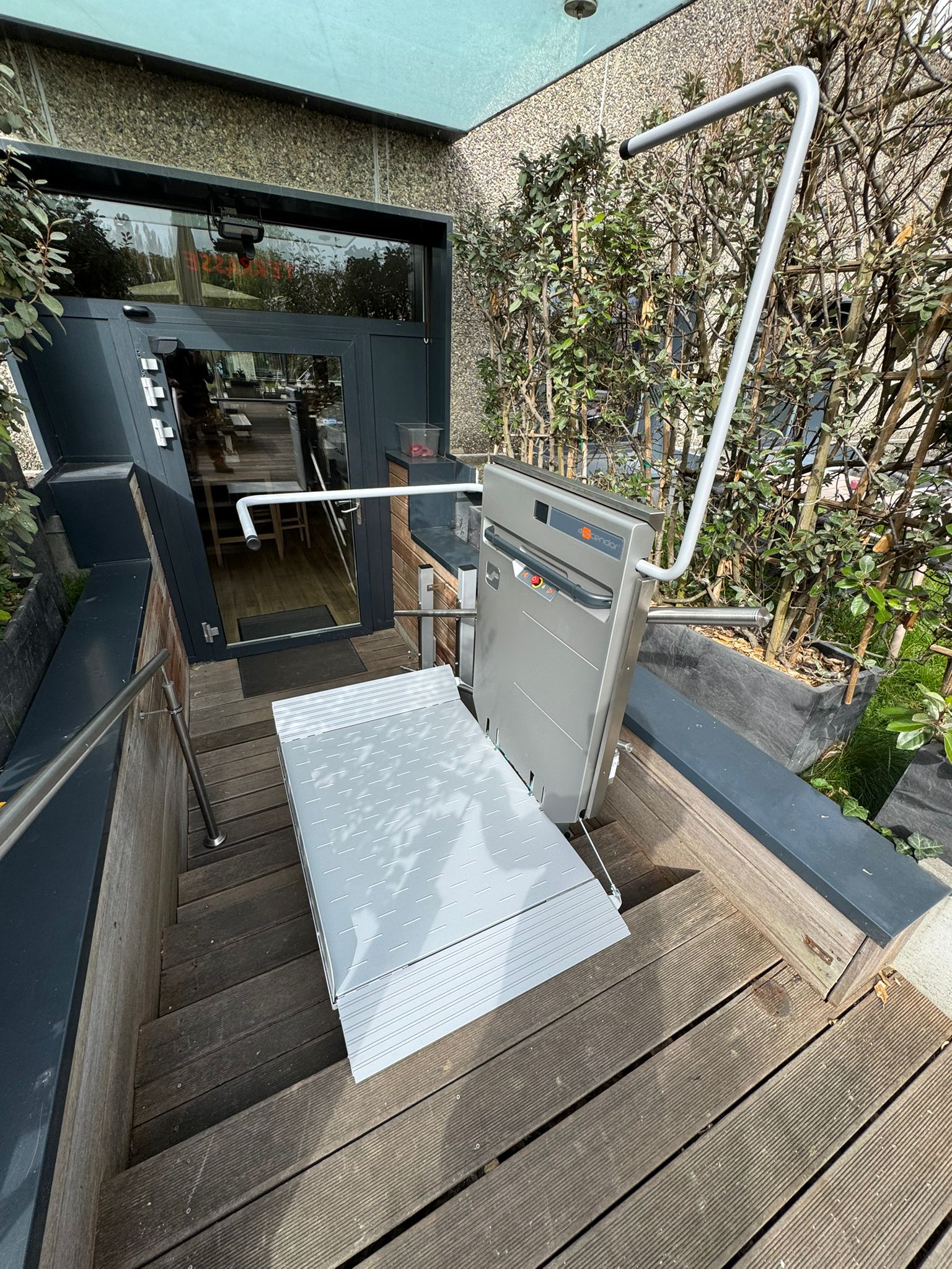 LSM Mobility Plateforme élévatrice PLG7 extérieure installée sur un escalier en bois menant à une porte d'entrée vitrée. La plateforme est grise avec une rampe abaissée, facilitant l'accès pour les personnes à mobilité réduite.