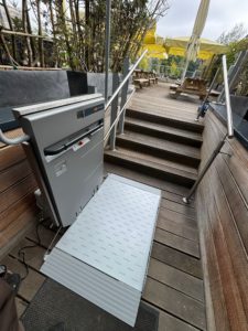 LSM Mobility Plateforme élévatrice PLG7 extérieure en acier inoxydable, installée près d'un escalier en bois menant à une terrasse avec des parasols jaunes. Accès PMR facilité.