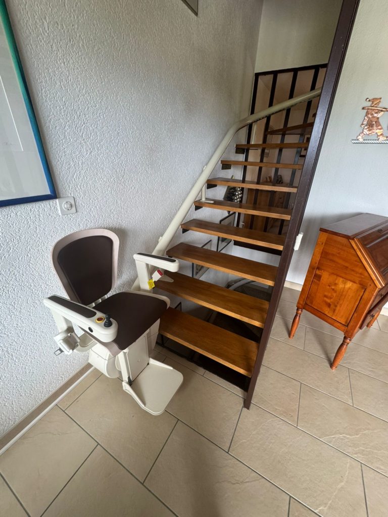 LSM Mobility Un siège monte-escalier beige et marron installé dans une villa, facilitant l'accès à l'étage. L'escalier en bois, avec sa rampe blanche, mène à un niveau supérieur. Meuble en bois visible à droite.