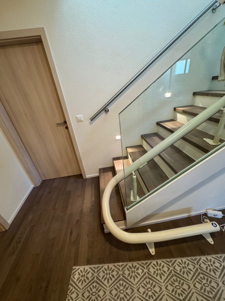 LSM Mobility Escalier moderne avec monte-escalier monorail courbe Handicare blanc. Marches en bois foncé, rampe en verre et acier, porte en bois clair à gauche. Tapis à motifs au sol.