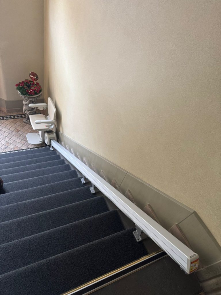 LSM Mobility Siège monte-escaliers Handicare 1100 replié sur un escalier intérieur avec moquette grise. Un pot de fleurs décore le palier.